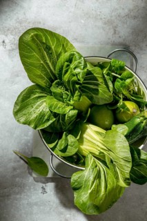 A colander filled with fresh green vegetables, including bok choy, lime, and zucchini, is placed on