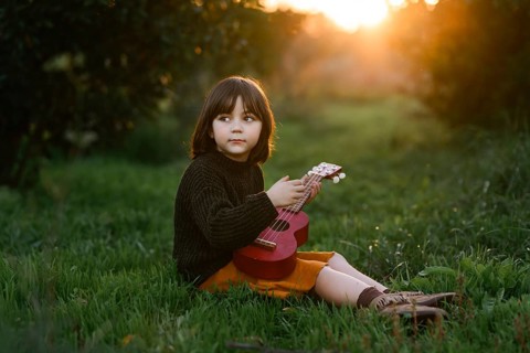 A young child sits on green grass with orange trees, holding a red ukulele, with the warm glow of