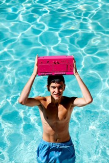 Smiling young teenage boy in blue swim shorts holds a vibrant pink radio above his head in a bright