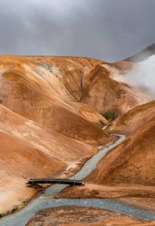 Steaming stream and bridge between colourful rhyolite mountains with snowfields, Hveradalir