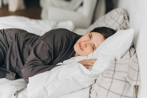 A young woman in comfortable loungewear lies on a white bed, resting her head on soft pillows while