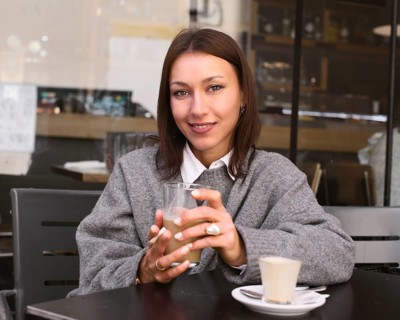 A woman in a gray sweater sits at a bar table, holding a cup of coffee. She smiles warmly, exuding