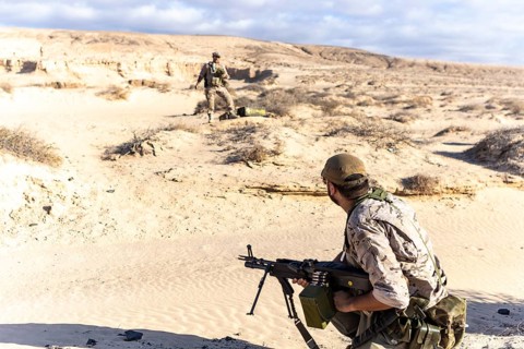 Two soldiers in camouflage gear conduct training exercises in a desert environment. One soldier is