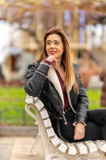Young woman wearing a black jacket is smiling while sitting on a bench in an amusement park