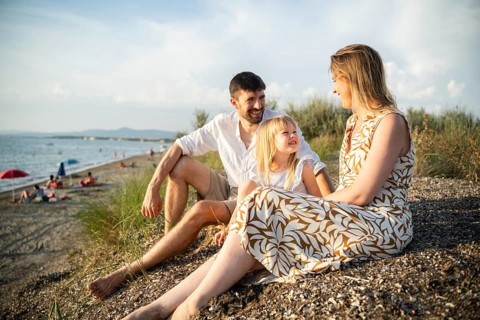 A young family sits on a sandy beach in Italy, enjoying a golden sunset. The parents lovingly