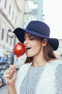 Elegantly dressed woman in a blue hat bites into a glossy candied apple. The urban backdrop