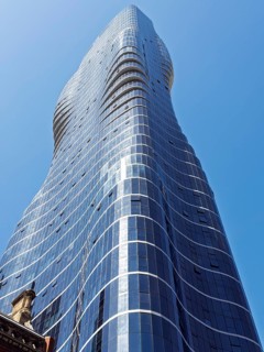 A modern skyscraper in Melbourne, Australia, rises dynamically against a cloudless blue sky. The