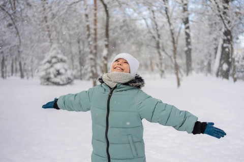 A child in a green coat, white hat, and scarf joyfully stretches their arms in a snow-covered park.