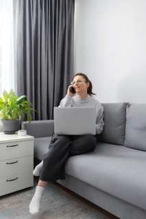 A woman sits on a sofa with a laptop and phone, multitasking in a cozy home office setting. Soft