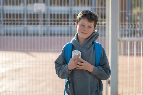 A young boy wearing a backpack and a hoodie holds his lunch while standing in front of the school