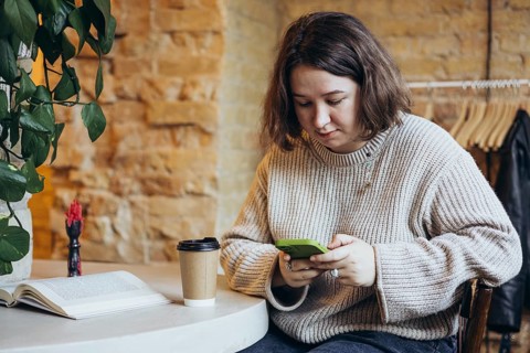 A young woman in a relaxed sweater sits in a cozy bookstore cafe, looking at her smartphone A