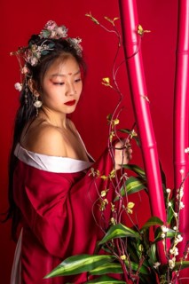 A woman in a red dress with a flower headpiece stands in front of a red background