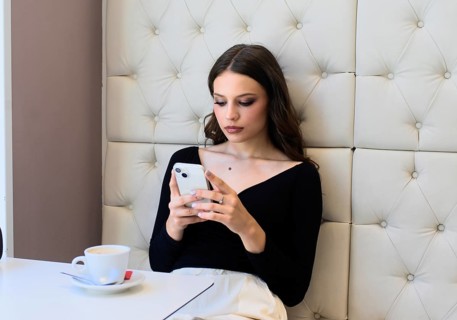 A woman in a cafe intently reads her smartphone. She sits against a plush, button-tufted wall, with