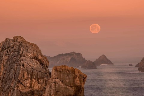 The full moon hovers above jagged rock formations along the Atlantic coast of Arnia Beach, Costa