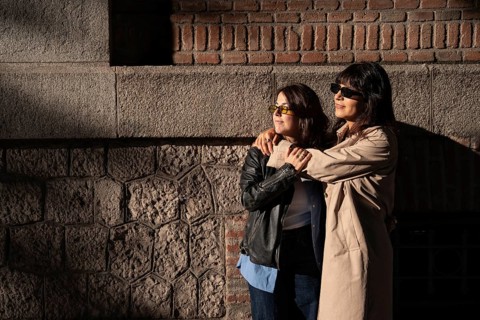 A multiethnic lesbian couple embraces warmly, basking in the sunlight against a brick and stone