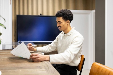 A young professional sits at a wooden desk, reviewing documents with a pen in hand. The modern