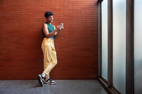 A person stands casually, texting on their phone against a red brick wall, wearing a green top and