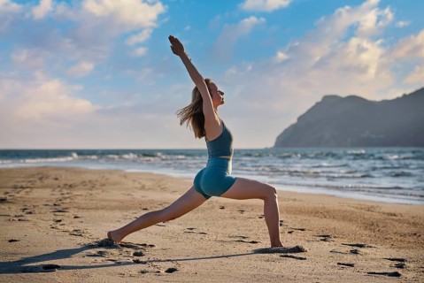 A woman is performing Virabhadrasana 1, or Warrior 1 pose, on the sandy beach as the sun sets. The