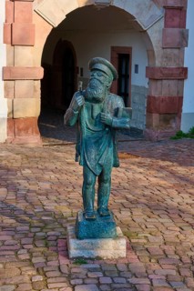 A bronze statue of a bearded man in front of a historic archway on a paved ground, Raibock, Market