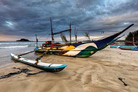An abandoned fishing boat is lying on an empty beach under a dramatic sky, the fishermen at Matara