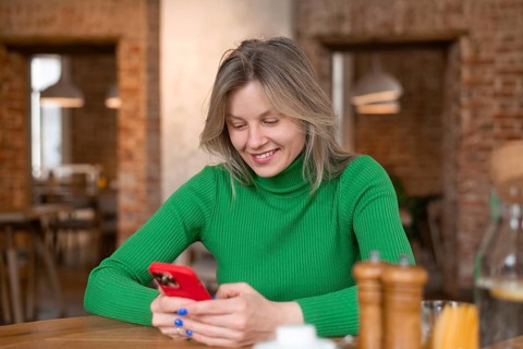 A woman in a green sweater smiles while using her phone at a cozy cafe. In the foreground, table