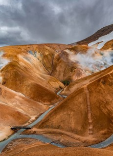 Bridge over steaming stream between colourful rhyolite mountains with snowfields, Hveradalir