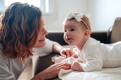 A joyful newborn baby girl wearing a headband smiles at her mother in a cozy, well-lit room. The