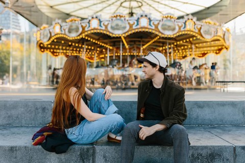 A young couple relaxes on concrete steps near a brightly lit carousel in Brooklyn Bridge Park. The