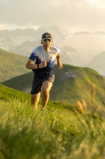 A determined man with sunglasses and a hat runs energetically through a lush, mountainous trail,