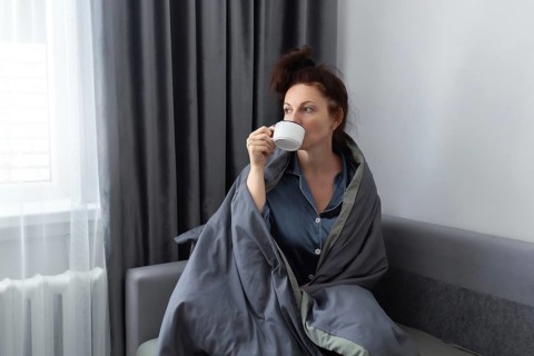 A woman in pajamas, wrapped in a gray blanket, enjoys her morning coffee on a sofa. Soft window