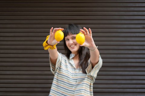 A smiling young woman holds two vibrant yellow lemons towards the camera, wearing a striped shirt