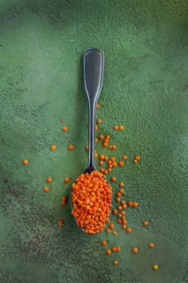 Bright orange lentils fill a silver spoon, with some lentils scattered on a green textured surface,
