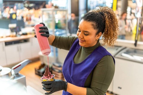 Smiling woman employee working in a food service business, wearing gloves and an apron, adding a