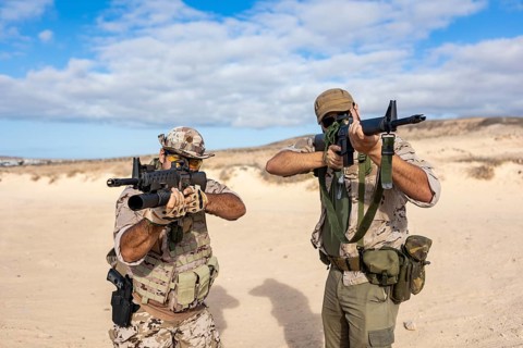 Two soldiers in tactical gear aim rifles while positioned in a desert environment. They wear