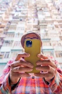 From below an unrecognizable man in a hat uses a smartphone in front of a dense Hong Kong