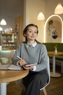 A woman sits in a cozy cafe, holding a tablet pencil and smiling, surrounded by modern decor. A