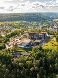 A large building surrounded by densely wooded hills and a cityscape, taken from the air under a