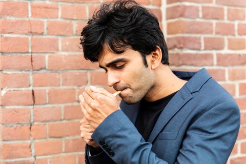 A indian man in a blue suit lights a cigarette while standing against a brick wall at a conference