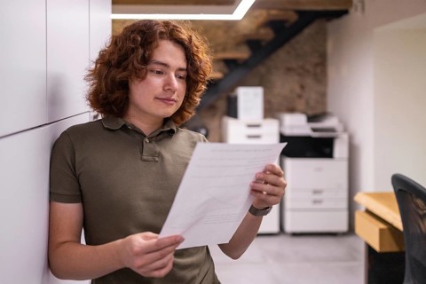 A young man with curly hair leans against a wall while thoughtfully reading a document in a cozy,