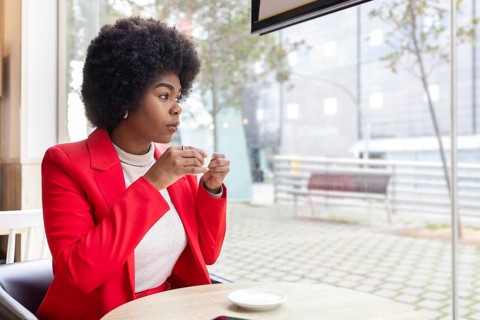 A woman in a vibrant red jacket enjoys a cup of coffee, seated in a modern cafe. The outdoors