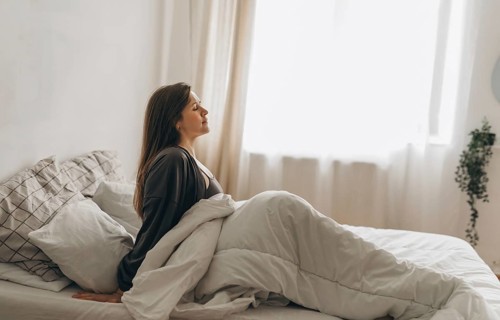 A young woman sits in bed with her eyes closed, wrapped in soft white blankets. She appears calm