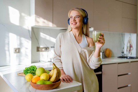 A woman with headphones enjoys a green smoothie in a sunlit kitchen. Fresh fruits and vegetables on
