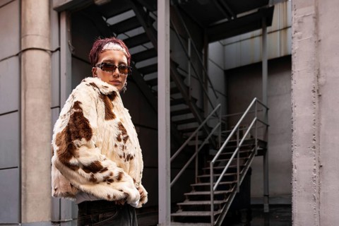 A stylish woman wearing a faux fur coat and sunglasses stands confidently in an urban alley. The