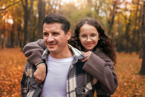 A father carries his daughter on his back in a forest during autumn. The ground is covered with