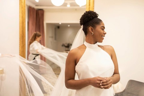 A bride smiles elegantly as she tries on a sophisticated wedding dress in a warmly lit bridal shop.