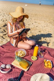 Woman in straw hat and striped shirt relaxes on maroon picnic blanket at the beach, using a tablet