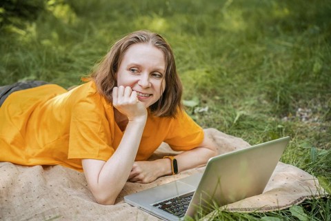 A woman in a bright yellow shirt lies on a blanket in a park, using a laptop. She appears relaxed
