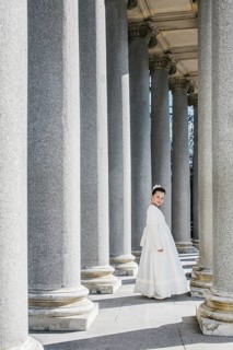 A young girl stands gracefully in a First Communion Day dress, surrounded by towering stone columns