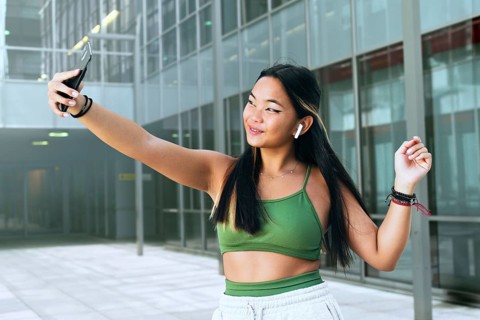A joyful teenager takes a selfie in a modern urban setting, wearing a green top and wireless