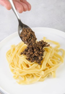 A hand elegantly spoons rich black truffle sauce onto a bed of freshly cooked tagliatelle pasta,